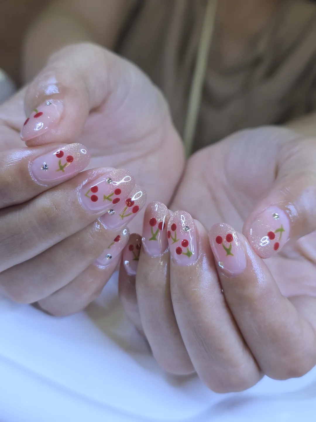 Hand with sheer pink almond nails painted with red cherries and small silver rhinestones, design A511 at Nanny Nails Nakhon Phanom